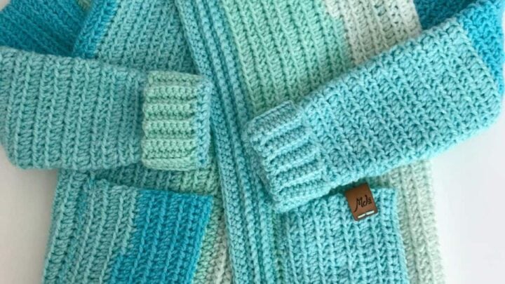 Hand-crocheted cardigan in blue, green, and white with two front pockets, textured stitches, and a button collar.
