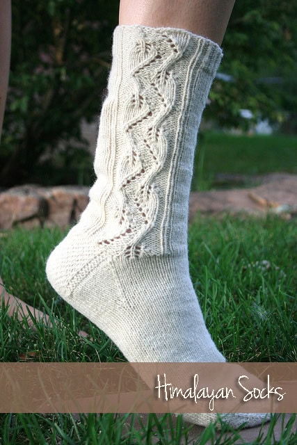 A person standing on grass wearing a hand-knitted sock with intricate cable patterns on the calf, labeled "Himalayan Socks." The background is softly blurred with greenery. -Marly Bird