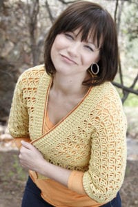 A person with short brown hair is outdoors, wearing a yellow crocheted cardigan over an orange top. The person is smiling slightly, with hands clasped in front. The background is blurred with trees and greenery. -Marly Bird