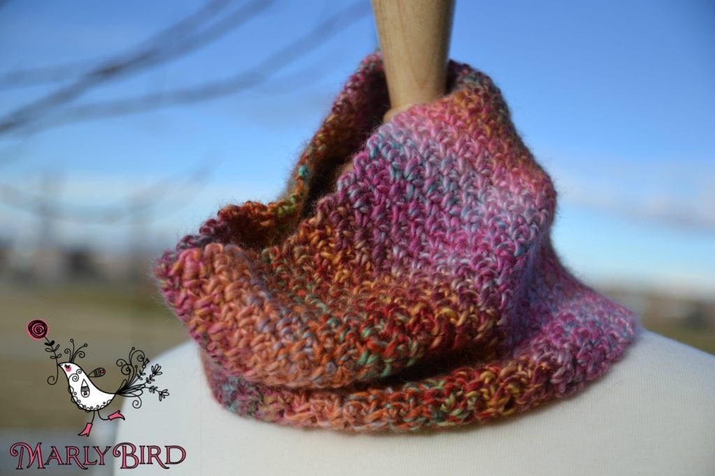 Close-up of a colorful knitted scarf wrapped around a mannequin's neck, with a blurred natural background, showcasing the detailed texture of the yarn. -Marly Bird