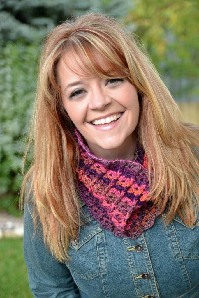 A joyful woman with shoulder-length blonde hair wearing a denim jacket and a colorful crocheted scarf, smiling in a grassy outdoor area with blurred greenery in the background. -Marly Bird