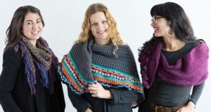 Three women standing together, smiling, wearing colorful knitted scarves. The woman in the middle holds a multi-colored crochet triangle shawl. They stand against a plain white background. -Marly Bird