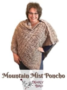 A smiling woman standing and modeling a beige crochet poncho called "Mountain Mist Poncho by Marly Bird". She has glasses and her hair styled up. -Marly Bird