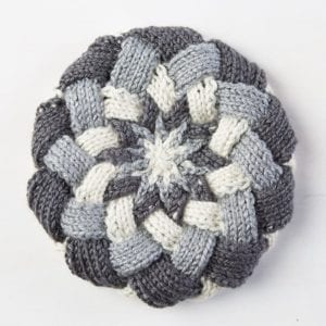 A handmade, circular knitted potholder featuring a starburst pattern in shades of gray and white yarn on a plain white background. -Marly Bird