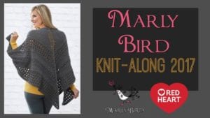 A woman models a gray textured triangle shawl, smiling over her shoulder, against a white brick wall. Text on the right reads "Marly Bird Knit-Along 2017" with the Red Heart yarn logo. -Marly Bird