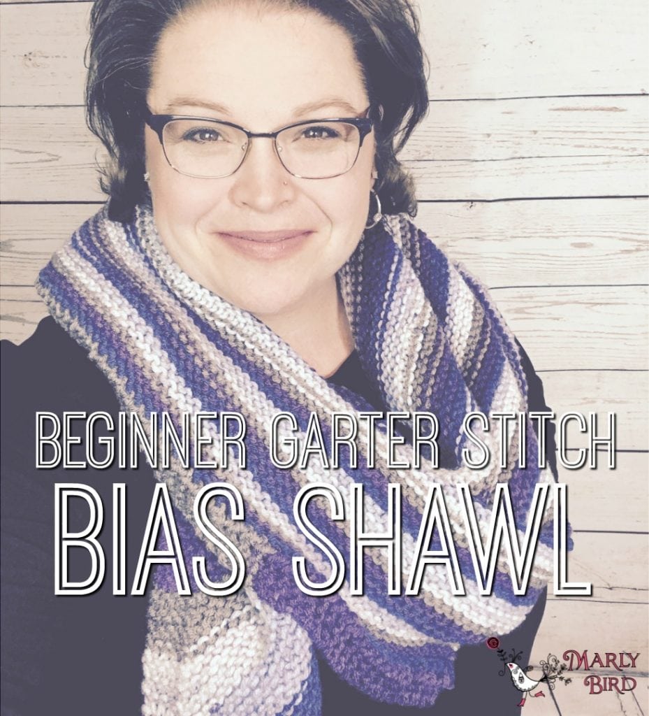 A smiling woman wearing glasses and a striped, knitted shawl stands against a wooden plank background. Text overlay reads "Beginner Garter Stitch Bias Shawl | BHooked Crochet Podcast. -Marly Bird