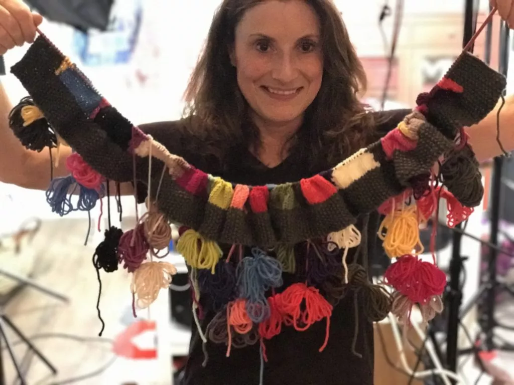 A woman smiles as she holds up a colorful Intarsia knit blanket with multiple strands of yarn hanging from it, standing in a room with soft lighting. -Marly Bird