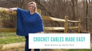 A woman stands smiling by a wooden fence in a park, wearing a blue Crochet Cables sweater. Text on the image reads "Crochet Cables Made Easy, Book Review by Marly Bird. -Marly Bird