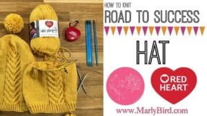 Knitting supplies and a yellow knitted hat on a wooden table next to a cover of a knitting video tutorial titled 'Road to Success Hat' by Marly Bird with the Red Heart brand logo. -Marly Bird