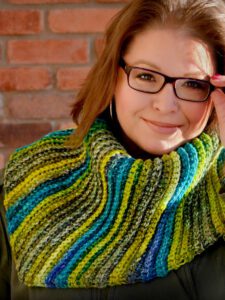 A smiling woman with glasses wearing a colorful crochet shawl against a brick wall background. -Marly Bird