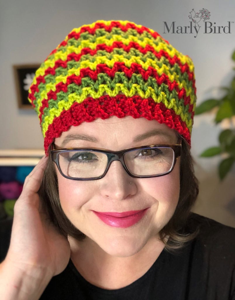Person models a red, green, and yellow crochet hat; close-up shows stitch detail and cozy yarn texture. Blurred home background.