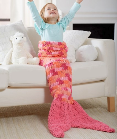 little girl in a mermaid tail blanket sitting on a couch with arms raised