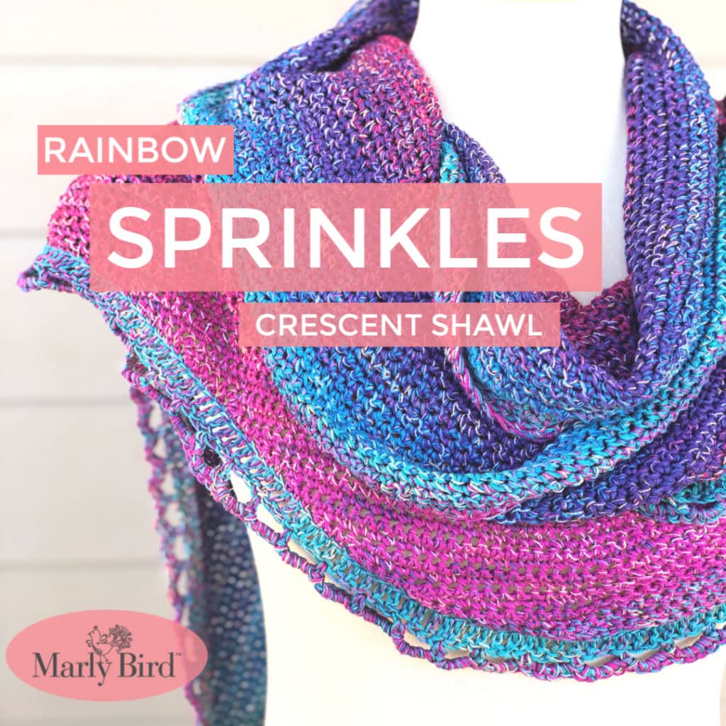 A crescent-shaped shawl knit in purple, blue, and pink yarn, shown draped on a white mannequin; Rainbow Sprinkles Shawl.