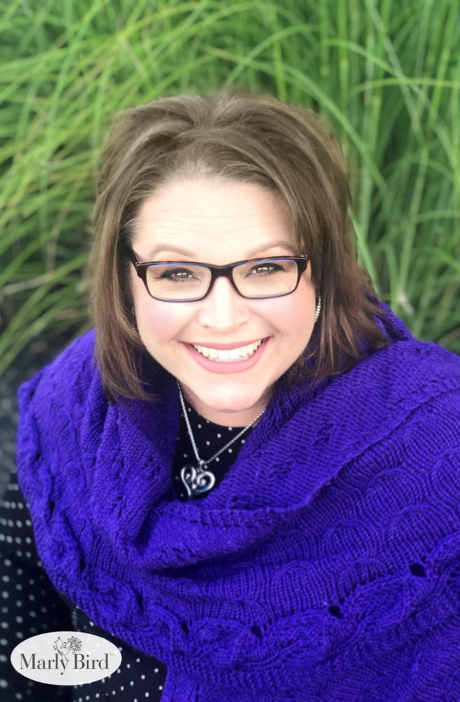 Woman models a vibrant purple crochet shawl with textured stitch detail, seated outdoors against tall green grass.