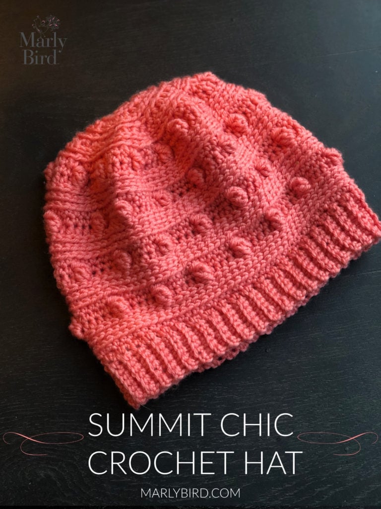 Coral crochet hat featuring bobble and ribbed stitches, shown flat on a dark background. Project: Summit Chic Crochet Hat.