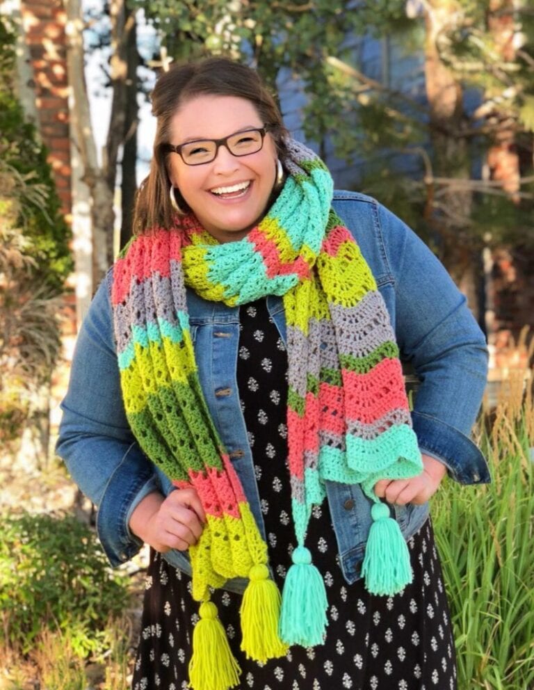 A joyful woman wearing glasses, a denim jacket, and a colorful Marly Bird knitted scarf smiles broadly in a sunny, outdoor setting with trees in the background. -Marly Bird
