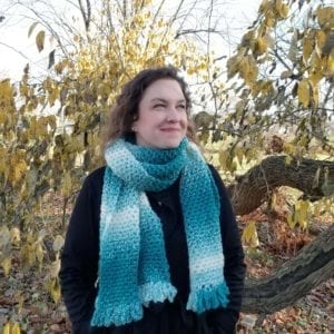 A woman wearing a black jacket and a turquoise-and-white striped scarf smiles, looking to the side in a wooded area with golden leaves, reminiscent of the cozy settings often featured on Moogly Blog. -Marly Bird