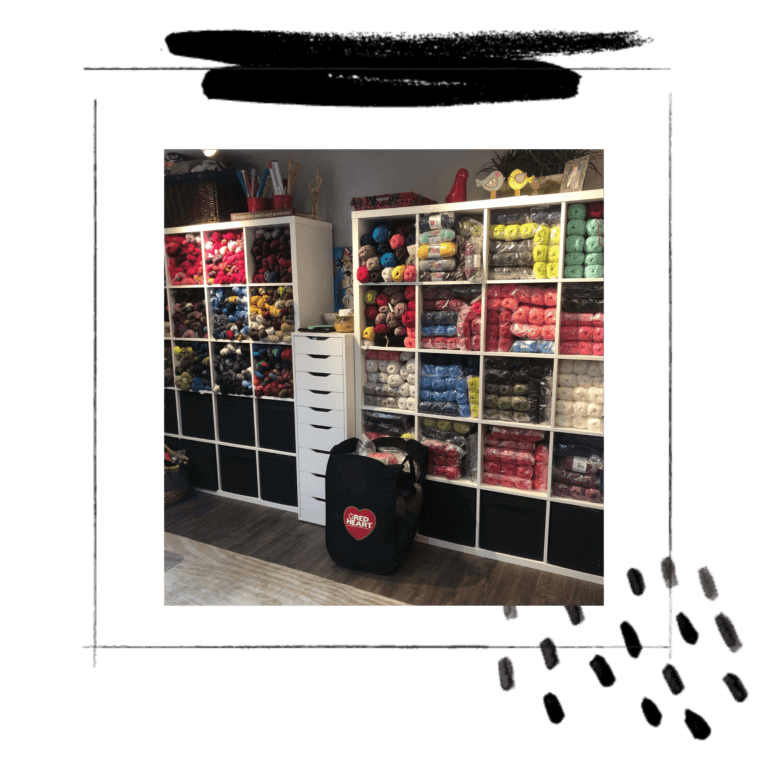 A well-organized yarn store with shelves full of colorful stash busting yarn balls. A black backpack with a logo sits on the floor next to a white storage unit. Footprints lead out of the scene. -Marly Bird