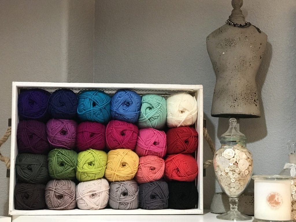 Colorful yarn balls in a white box, jar of buttons, candle, and dress form displayed on white shelf for crafting organization.