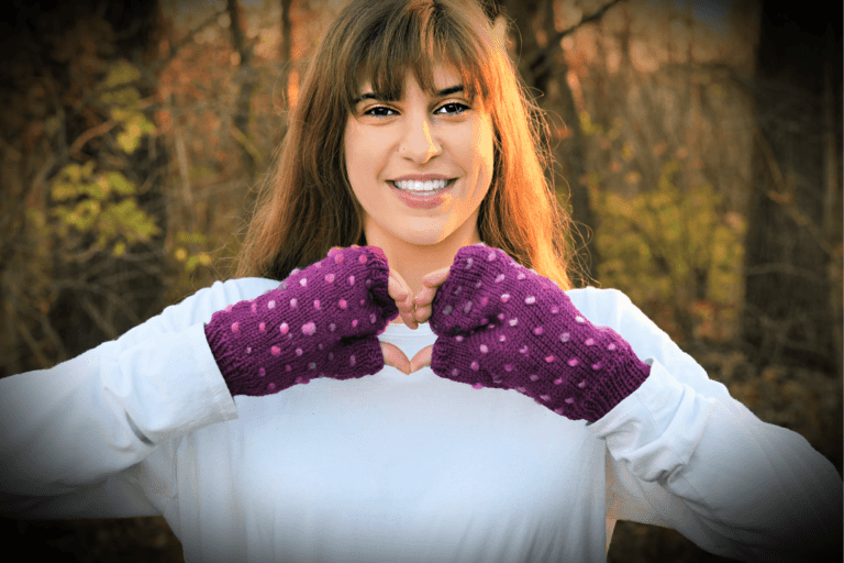 A person with long brown hair and bangs is smiling, standing outdoors in a forest. They are wearing a white top and purple fingerless gloves with pink polka dots, perfect for stash busting those small bits of yarn. They're forming a heart shape with their hands against the backdrop of autumn-colored trees. -Marly Bird