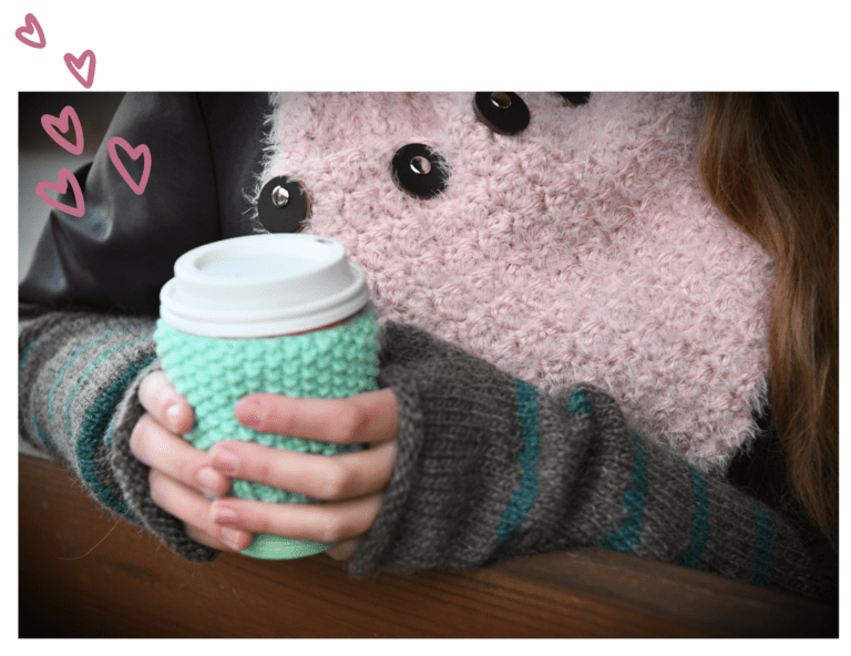 A person in a gray sweater clutching a coffee cup with a green cozy, partially obscured by a large pink plush with button eyes, Stash Busting texture and doodled hearts visible. -Marly Bird