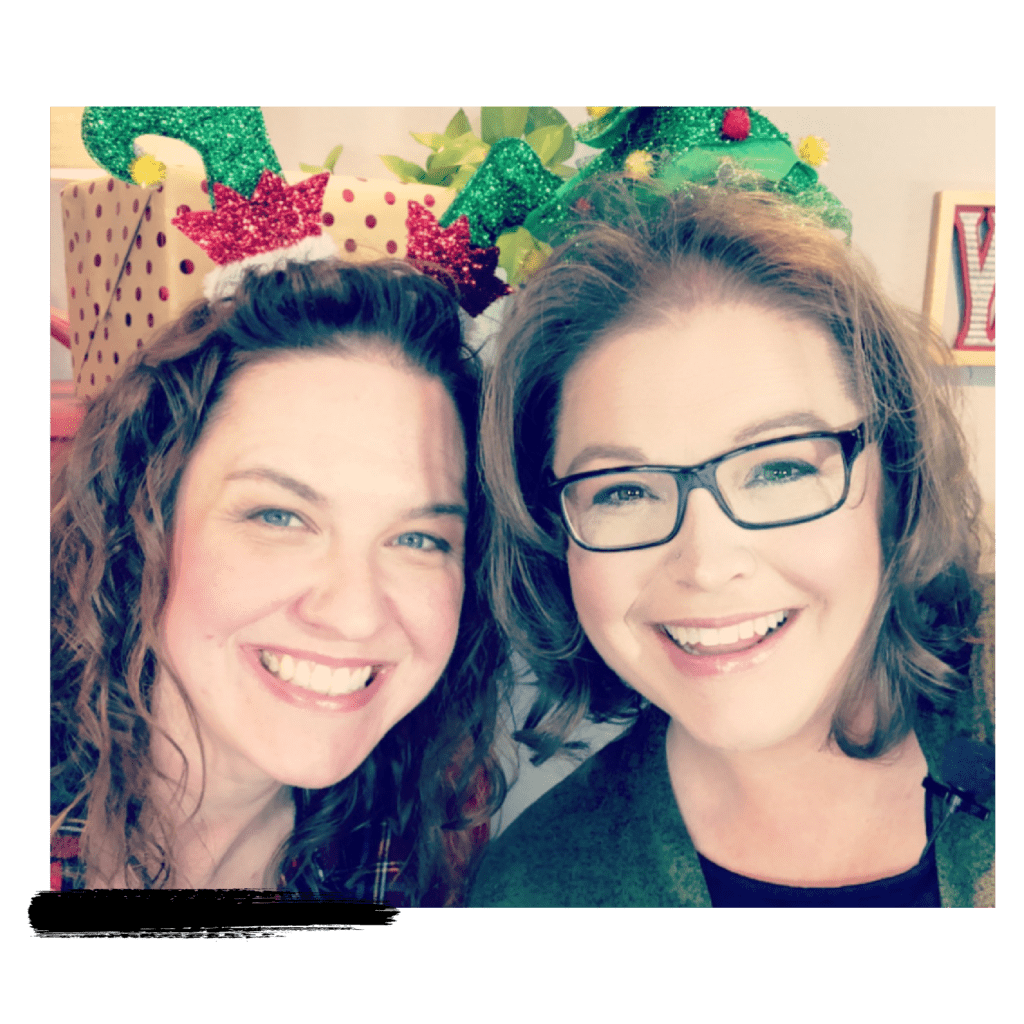 Two women smiling at the camera, one wearing glasses. They both have festive hats crocheted with glittery Christmas motifs like holly and reindeer antlers. -Marly Bird