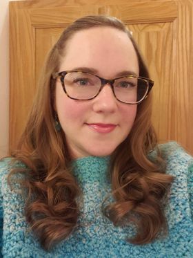 A woman with glasses, wearing a blue sweater featuring Marly Bird's knit and crochet patterns, smiles gently at the camera with a wooden door in the background. Her brown hair is styled in loose curls. -Marly Bird