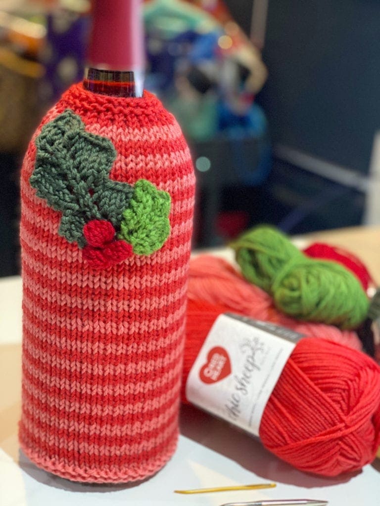 Knitted wine cozy in red and pink stripes with crocheted holly embellishment; yarn balls and hook beside the project.