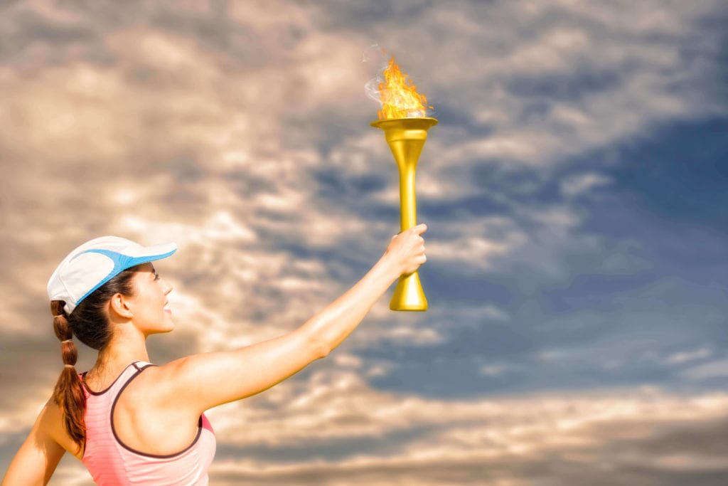 A woman in a cap and tank top holds up a colorful flaming torch against a dramatic sky with clouds. -Marly Bird