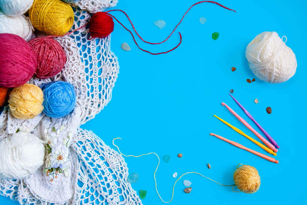 Colorful yarn balls and crochet hooks on a blue background, with a lace fabric and decorative beads, crafting supplies for the Classic Crochet Sock Workshop. -Marly Bird