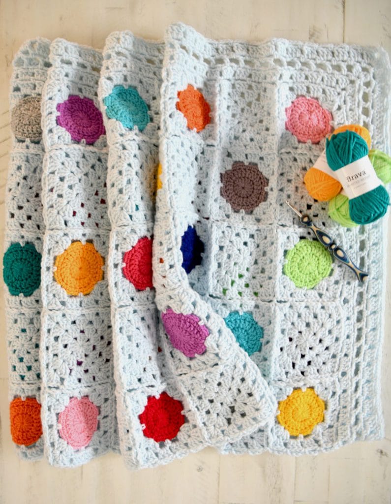 Crocheted blanket with white base and colorful circular motifs, folded beside orange, turquoise, and yellow yarn balls and crochet hook.