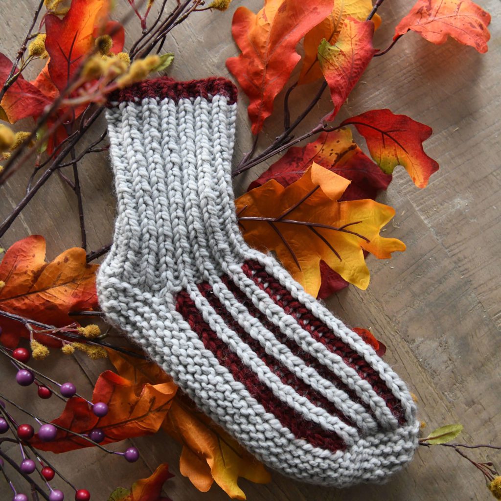 A knitted sock in gray and burgundy laid on a wooden surface surrounded by colorful autumn leaves and purple berries, showcasing a Stash Busting pattern. -Marly Bird