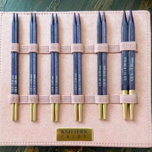 A set of six blue knitting needles organized neatly in three pink elastic bands on a pink holder labeled "J'Adore Gift Set." Each needle has its size imprinted in white. -Marly Bird