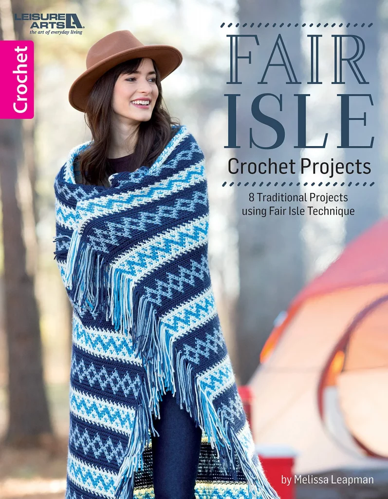 Front cover of the Fair Isle Crochet Projects Book, by Melissa Leapman, showing model wrapped in fringed, striped, and zigzag patterned blanket in 3 shades of blue and white. 