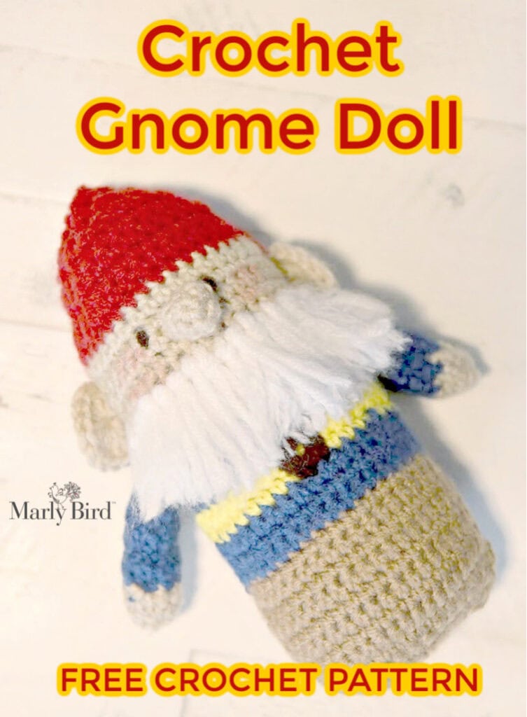 Image of a crochet gnome doll with a red hat, white beard, and a multicolored body displayed against a white wooden background. The text above reads "crochet gnome doll" and "free crochet pattern" by Marly Bird. -Marly Bird