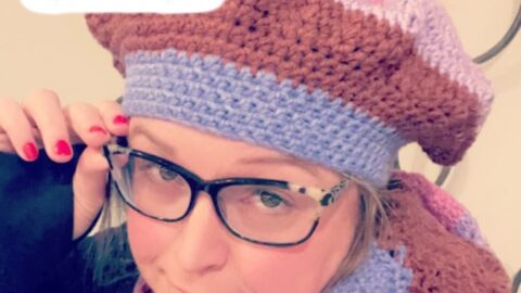 Glasses-wearing woman models a crocheted bonnet and scarf in purple, brown, and pink using textured Tunisian crochet stitches.