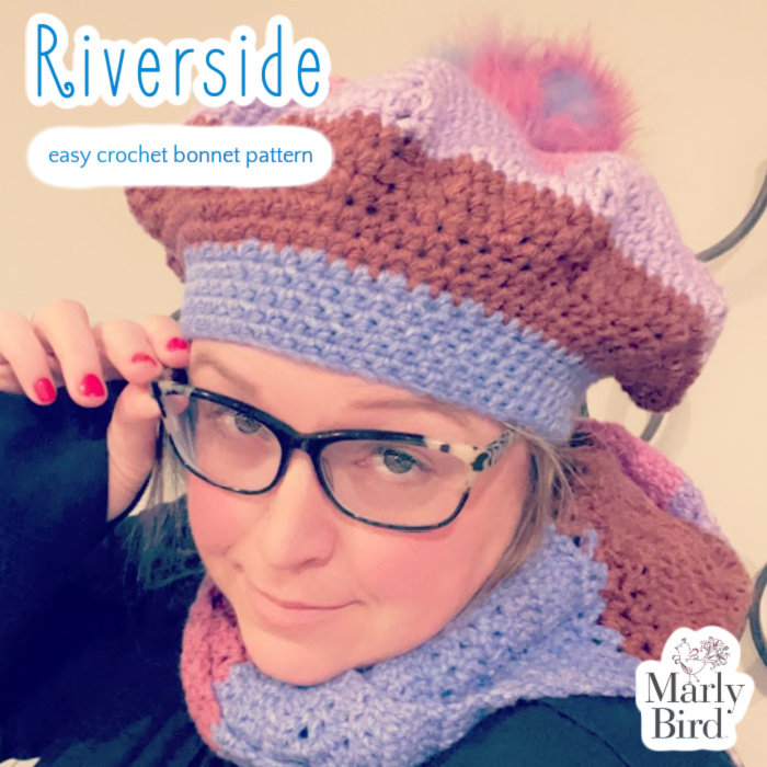 A woman with glasses wearing a handmade easy crochet bonnet with purple, blue, and brown hues, and a pink pompom, posing with a slight smile. Text overlay reads "Riverside - Easy Crochet Bonnet Pattern. -Marly Bird