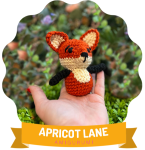 A hand holding a small, handmade crochet amigurumi fox named Felix against a backdrop of green plants. The fox is orange and white with black details, highlighted by a circular, stylized frame that reads "Apricot Lane Amigurumi. -Marly Bird