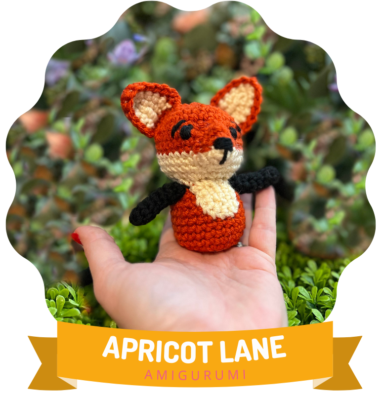 Felix the Fox in the Worsted weight yarn size. he is resting on the palm of marly's hand. the image is in a circle over a banner that reads Apricot Lane Amigurumi