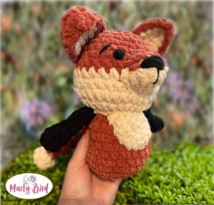 Hand holds a crocheted amigurumi fox with white belly, black limbs, brown body, large ears, and a black nose; leafy background.