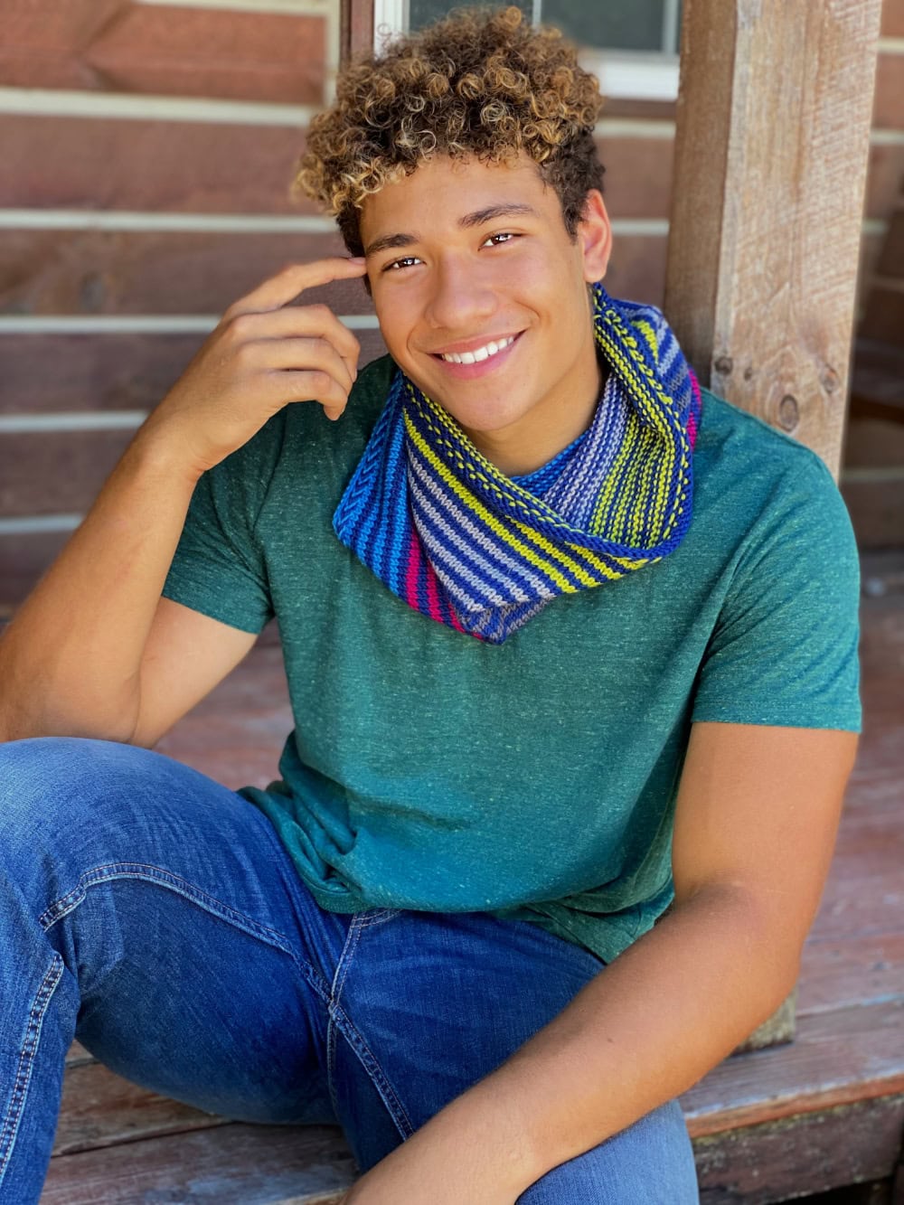 Bright knit cowl scarf with blue, yellow, pink, and white stripes drapes over a young man's teal tee and jeans.