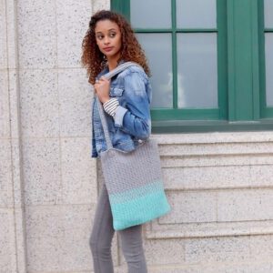 A young woman with curly hair facing sideways, holding a denim jacket over her shoulder. She carries a large Easy Split Crochet Tote, standing in front of a building with green windows. -Marly Bird