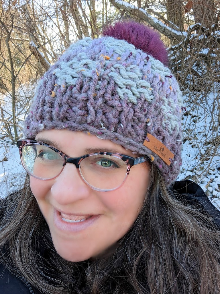 A smiling woman wearing glasses and a free Tunisian crochet hat with a purple pompom, enjoying a snowy backdrop. -Marly Bird