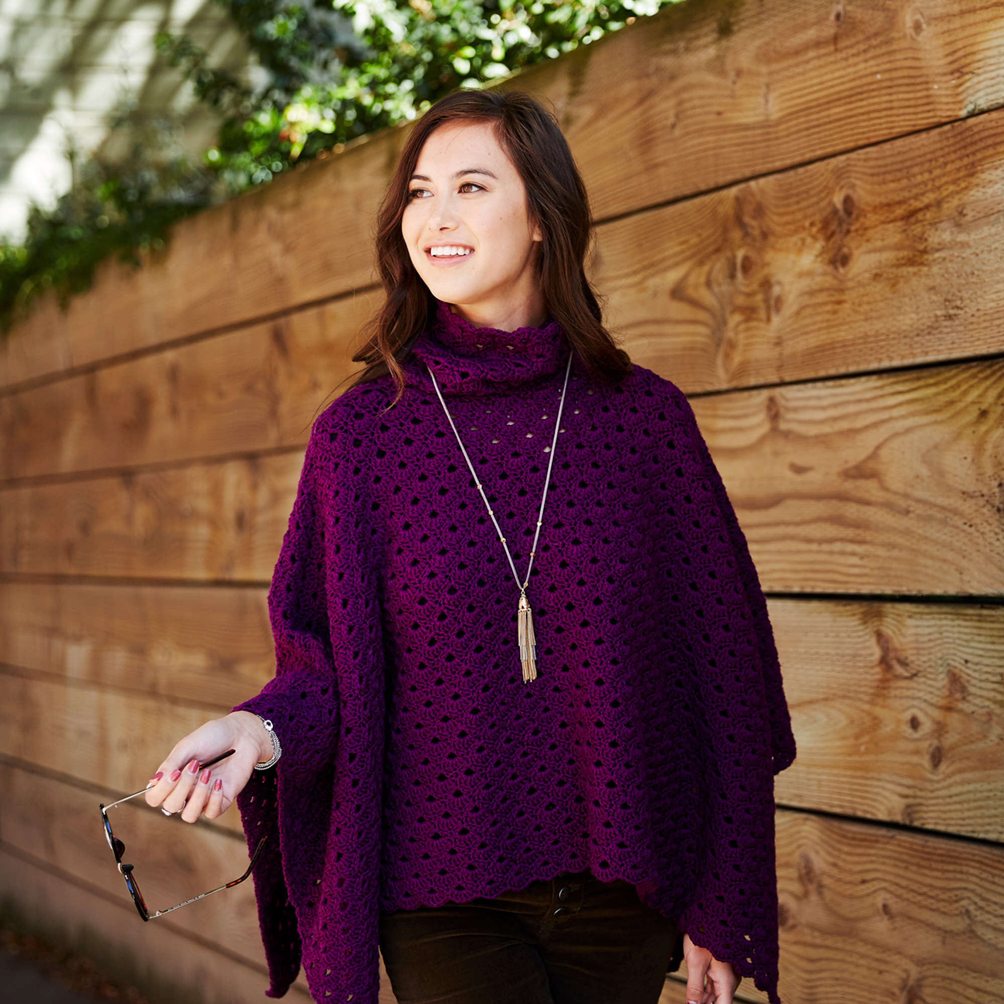 A woman with a cheerful expression stands outdoors, wearing a purple easy crochet poncho and holding sunglasses. She gazes to the side against a wooden fence backdrop. -Marly Bird