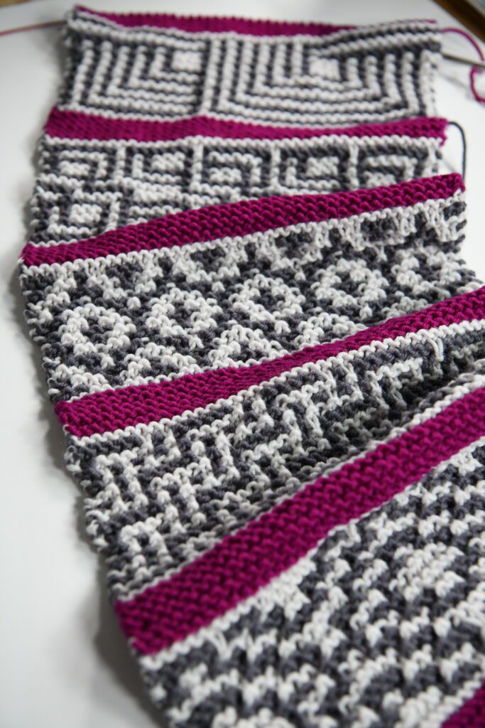 Close-up of a hand-knitted scarf in progress, featuring alternating stripes of gray, white, and vibrant pink yarn with various mosaic knitting patterns. -Marly Bird