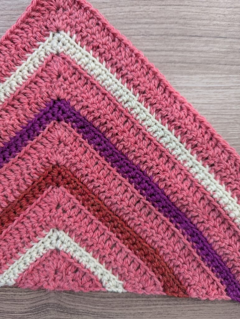 Close-up of a crocheted triangle in pink, white, red, and purple yarn showing neat rows and distinct stitch texture on wood surface.