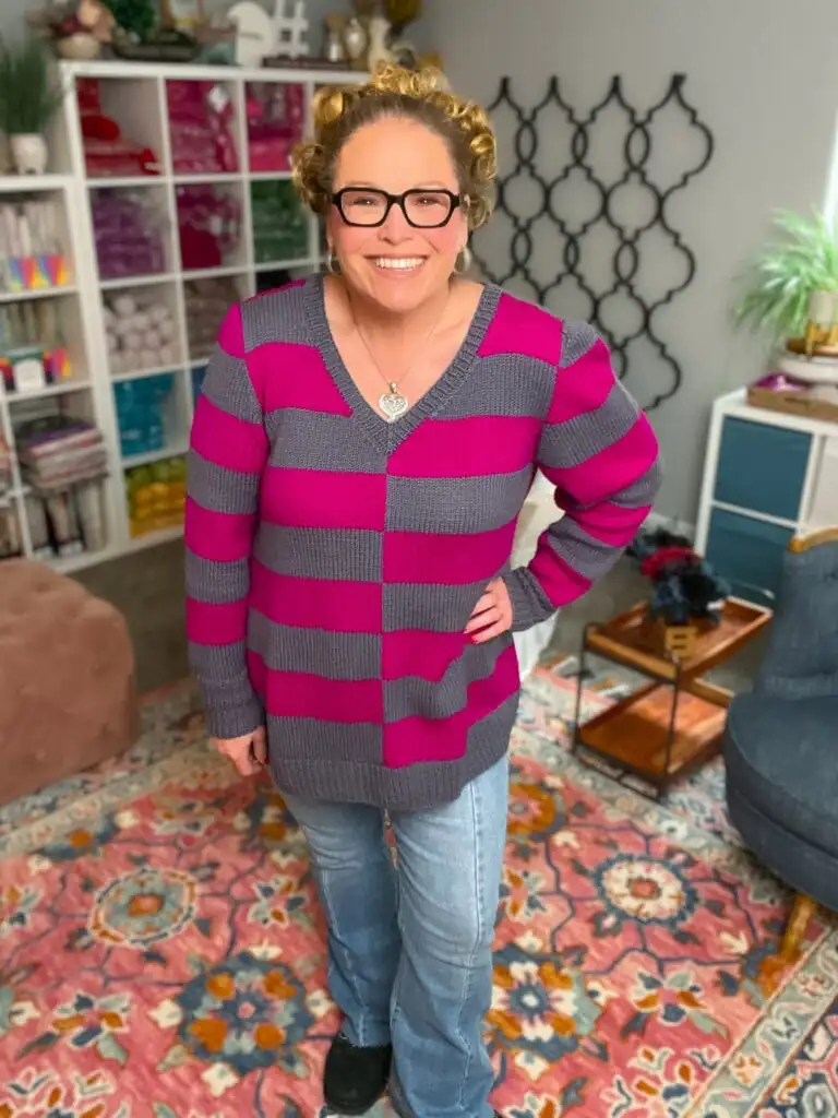 A smiling woman with curly hair, glasses, and a heart necklace stands indoors wearing her favorite knit sweater pattern in purple and magenta stripes, paired with jeans and black shoes. Behind her are bookshelves, decorative items, and a colorful rug. -Marly Bird