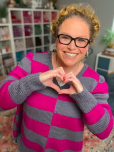 Woman in a gray and pink striped knit sweater, showing heart hands, stands by shelves with colorful yarn and crochet supplies.