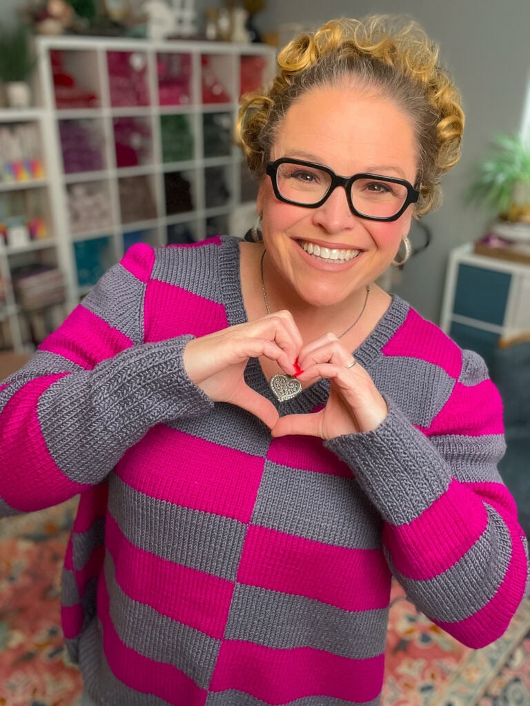 A smiling person with curly hair and glasses wears their favorite knit sweater pattern—a pink and gray striped sweater—and forms a heart shape with their hands. They stand indoors in front of shelves filled with yarn and colorful items. -Marly Bird