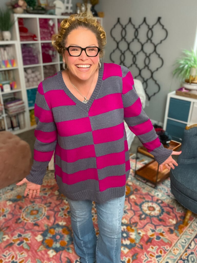 A smiling woman with curly blonde hair and glasses wears her favorite knit sweater pattern—a pink and gray striped sweater—with light blue jeans. She stands on a colorful patterned rug in a cozy room with bookshelves, plants, and wall art. -Marly Bird
